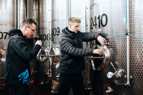 Two men checking the quality of red wine from a steel tank